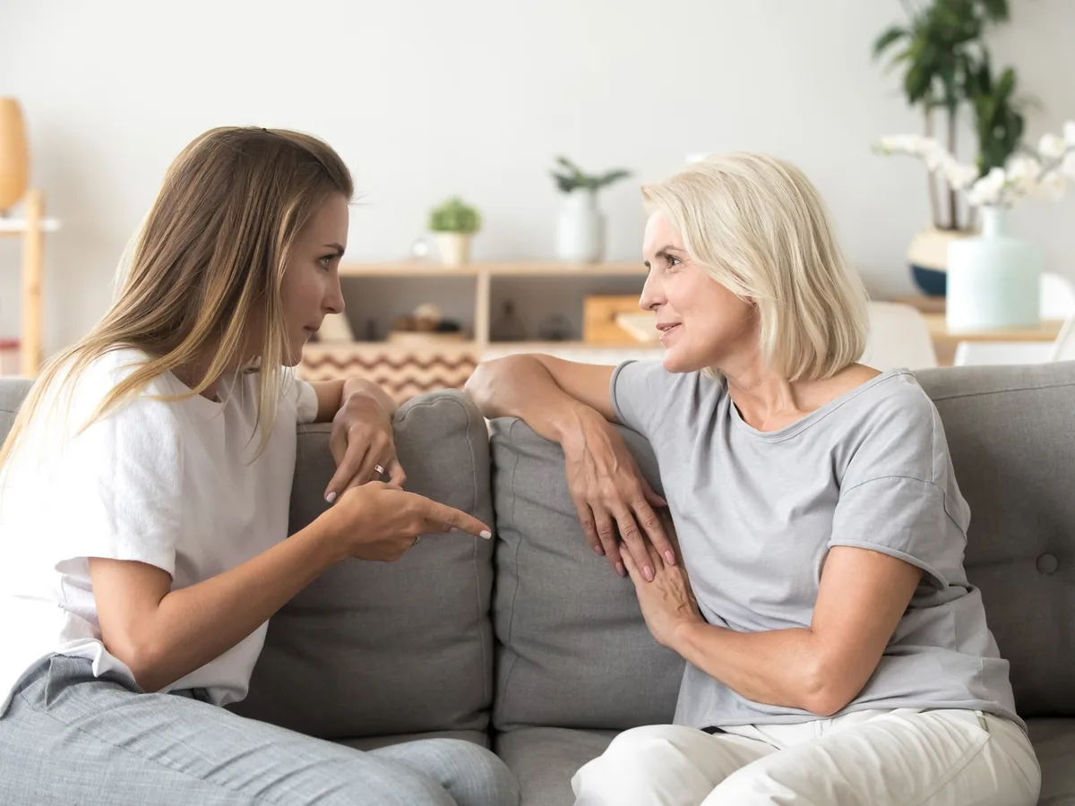 Two women in quiet conversation, sitting side by side on a sofa in a bright living room